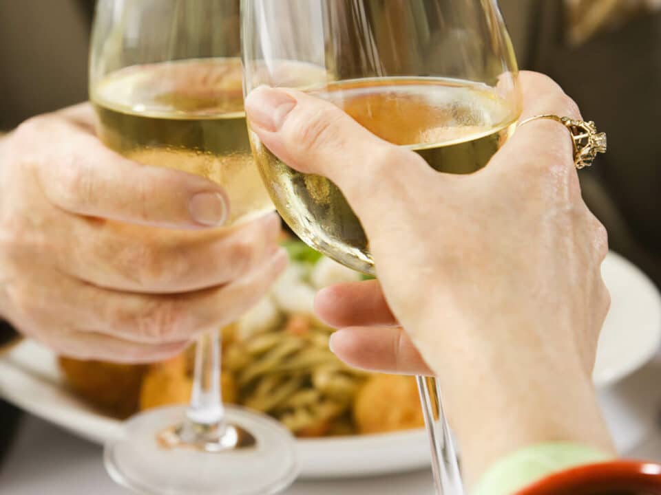 hands holding white wine glasses over a plate of food