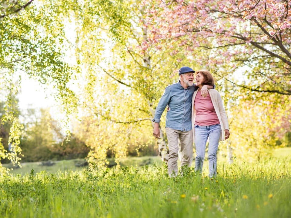 older couple walking through a field of blooming trees and grasses, with their arms around eachother