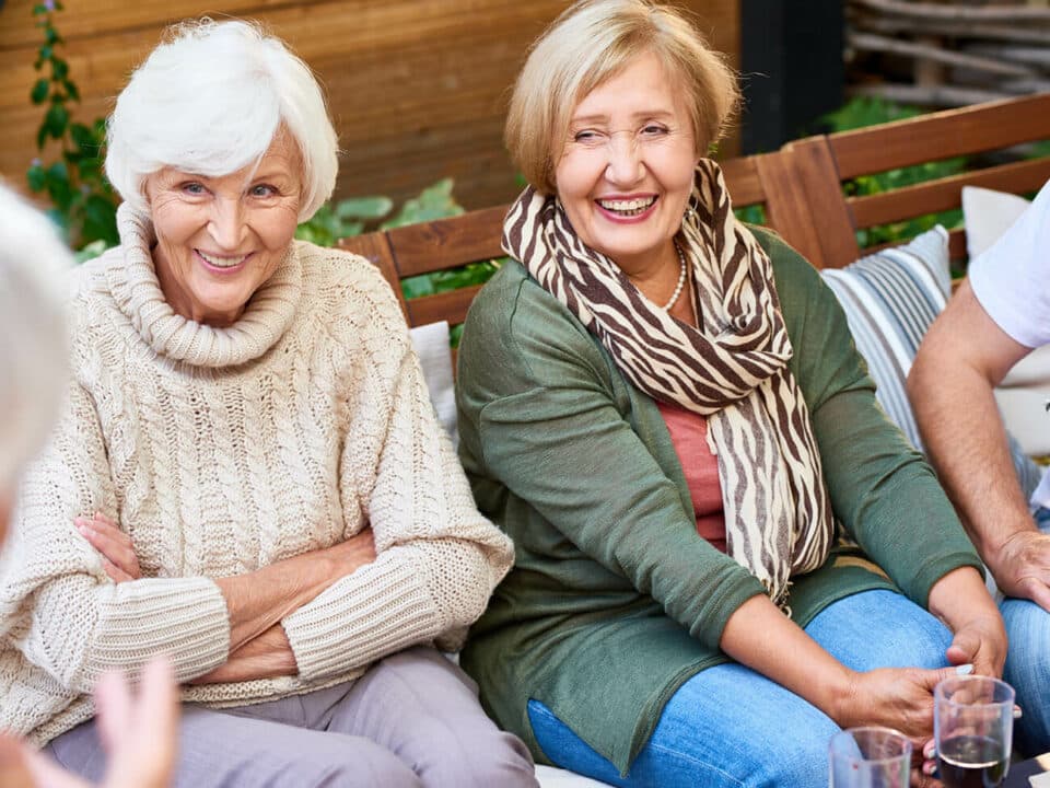 two women smiling, sitting on a bench, talking to another woman across from them with wine