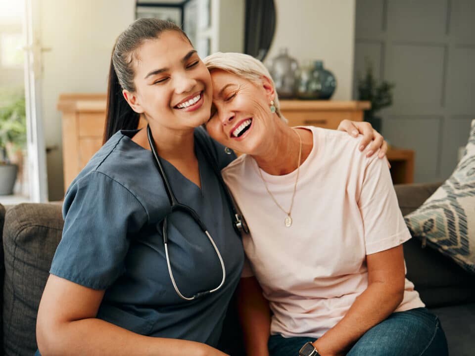 female caregiver with stethoscope hugging smiling woman in pink shirt