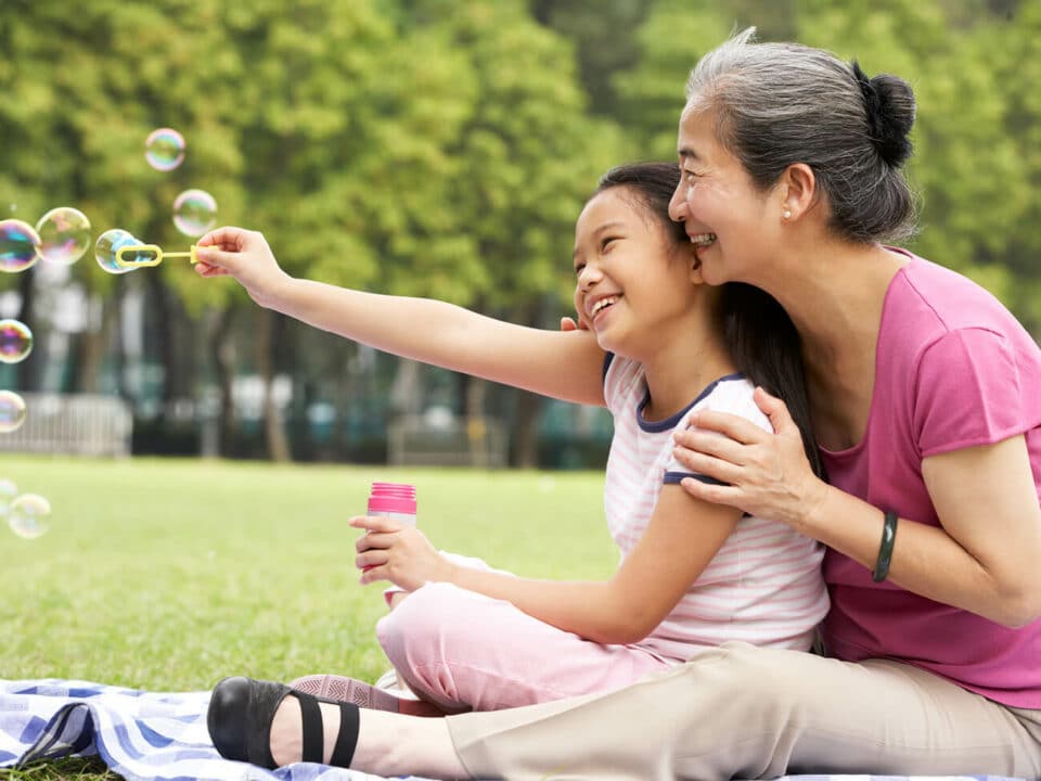 asian woman in pink sitting in a park with young girl blowing bubbles