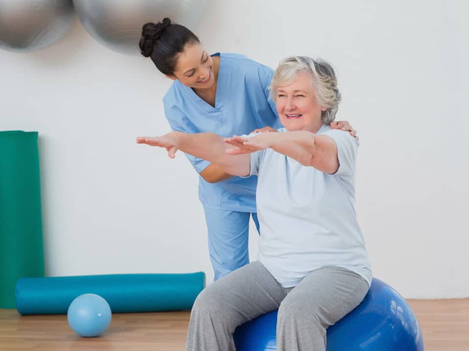 younger female caregiver assisting smiling woman on an exercise ball extending her arms