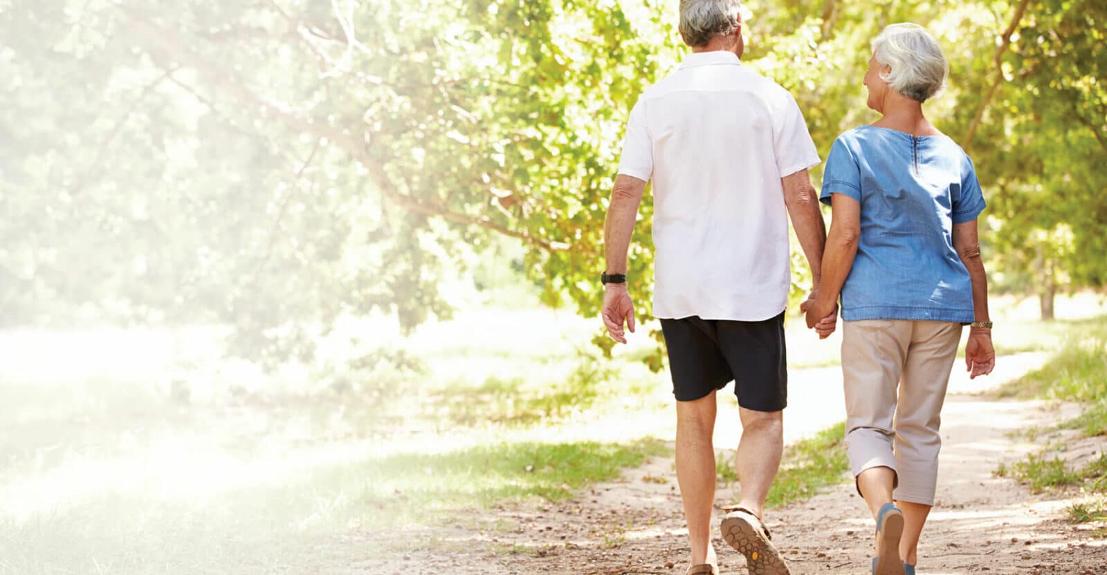 man in a white shirt and black shorts holding hands and walking with senior woman on a nature path
