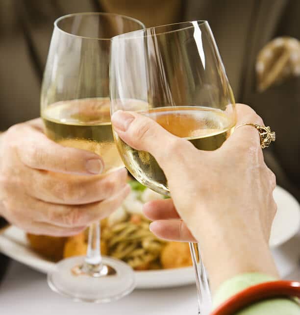 hands holding white wine glasses over a plate of food