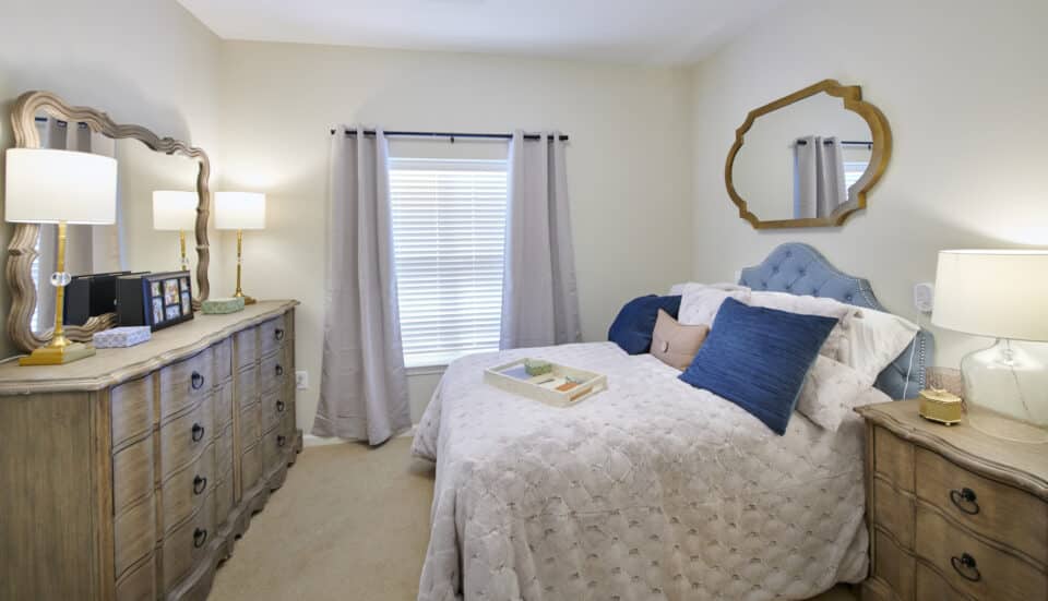 bed with blue upholstered headboard, pillows, dresser with two lamps and side table
