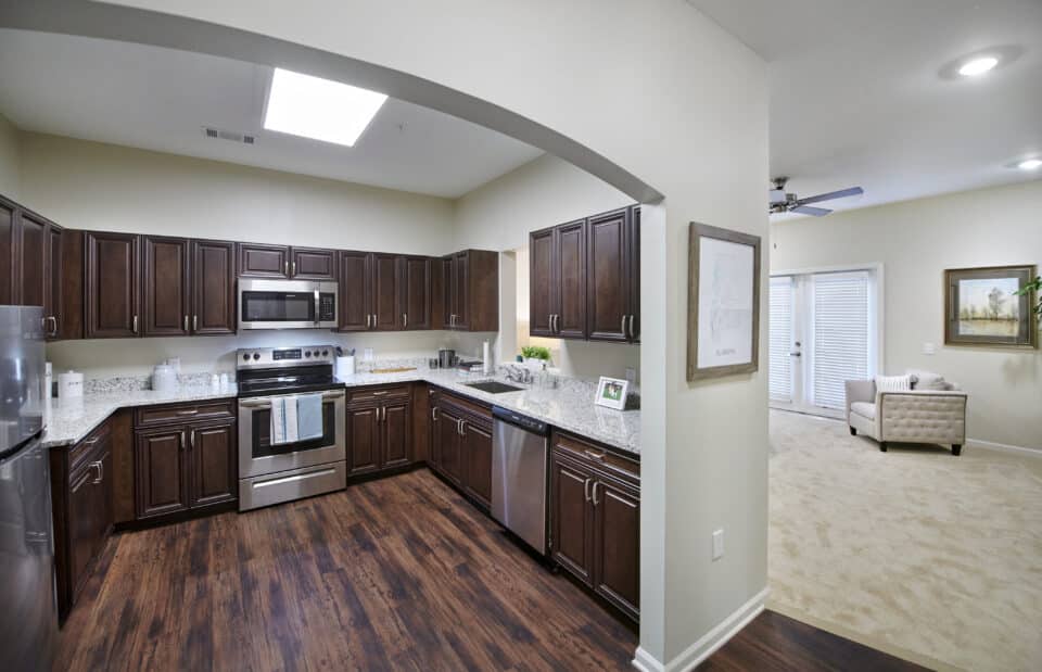 arched way kitchen, with brown cabinetry, stainless appliances, carpeted room adjacent with white upholstered chair
